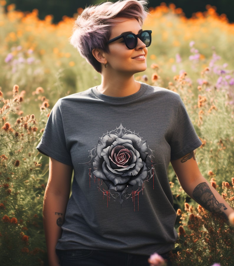 May include: A grey t-shirt featuring a graphic of a black rose with red dripping paint. The rose is surrounded by a decorative design.