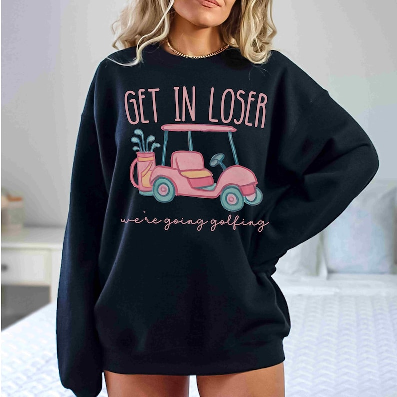 Puede incluir: Sudadera negra con un gr&aacute;fico de carrito de golf rosa y el texto "GET IN LOSER" arriba y "we're going golfing" abajo. El carrito de golf tiene ruedas azules y un asiento rosa. Estilo casual.