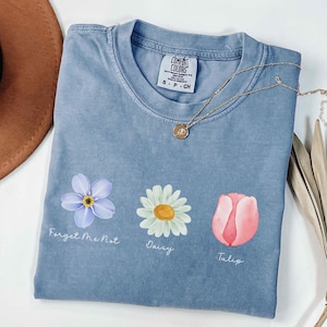 May include: A light blue t-shirt with a forget-me-not, daisy, and tulip design. The shirt has the words "Forget Me Not", "Daisy", and "Tulip" printed below each flower.