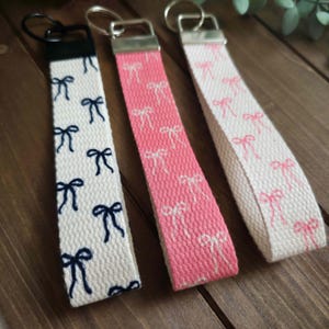 May include: Three keychains with fabric straps. One is white with navy blue bows, another is pink with white bows, and the third is white with pink bows. Each keychain has a metal ring for keys.