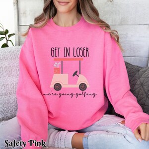 K&ouml;nnte beinhalten: Ein rosa Sweatshirt mit dem Text "Get in loser we're going golfing" und einem Cartoon-Golfwagen.