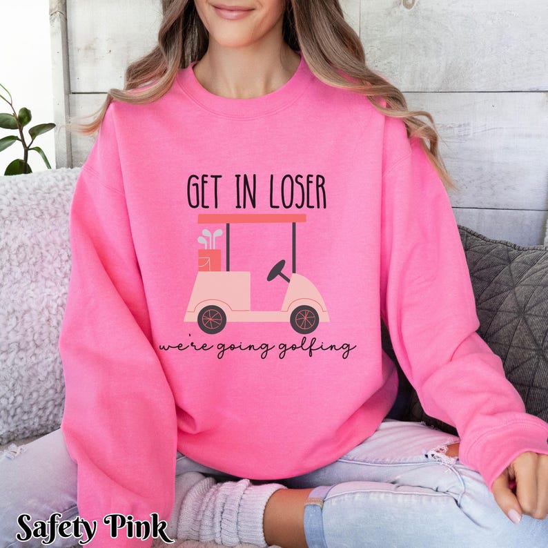 K&ouml;nnte beinhalten: Ein rosa Sweatshirt mit dem Text "Get in loser we're going golfing" und einem Cartoon-Golfwagen.