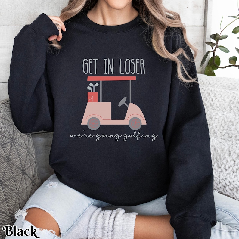 K&ouml;nnte beinhalten: Schwarzer Pullover mit dem Text "Get in loser, we're going golfing" und einer pinkfarbenen Golfwagen-Grafik.