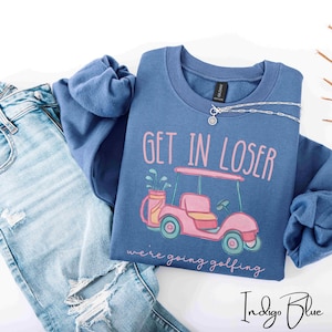 Puede incluir: Sudadera azul &iacute;ndigo con el texto "GET IN LOSER" sobre un gr&aacute;fico de carrito de golf rosa. La frase "we're going golfing" est&aacute; debajo del gr&aacute;fico. Un collar plateado est&aacute; sobre la sudadera. Vaqueros azul claro en la imagen.