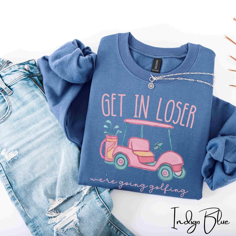 Puede incluir: Sudadera azul &iacute;ndigo con el texto "GET IN LOSER" sobre un gr&aacute;fico de carrito de golf rosa. La frase "we're going golfing" est&aacute; debajo del gr&aacute;fico. Un collar plateado est&aacute; sobre la sudadera. Vaqueros azul claro en la imagen.