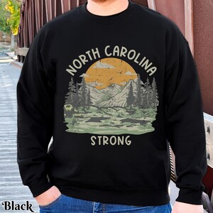 Op de afbeelding: Zwart sweatshirt met een grafisch ontwerp van een berglandschap en de tekst "North Carolina Strong".