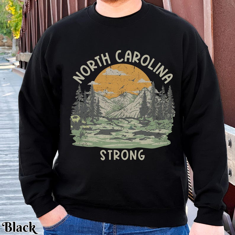 Op de afbeelding: Zwart sweatshirt met een grafisch ontwerp van een berglandschap en de tekst "North Carolina Strong".