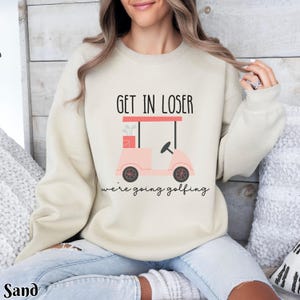 K&ouml;nnte beinhalten: Ein cremefarbener Pullover mit dem Text "Get in loser we're going golfing" und einer pinkfarbenen Golfwagen-Grafik.