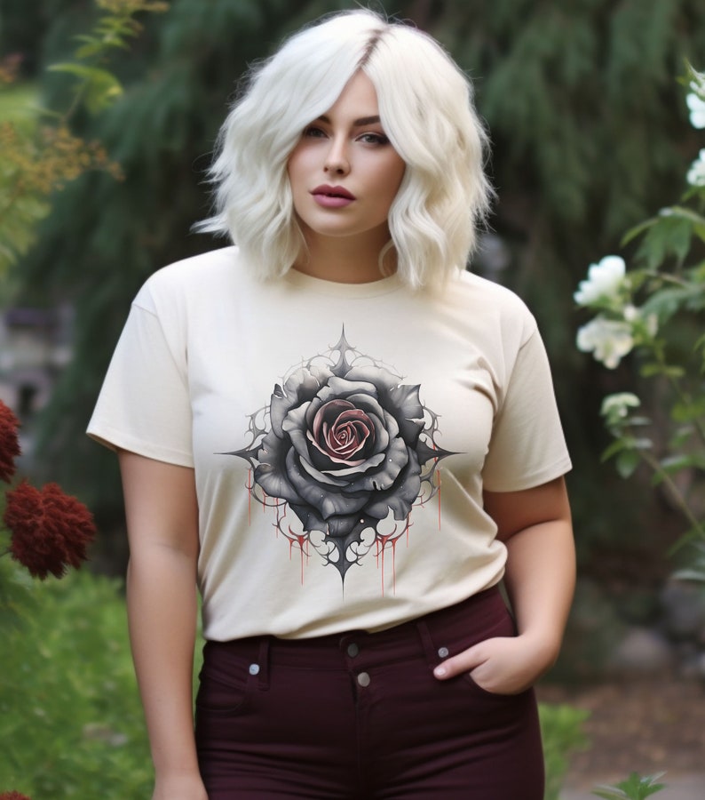 May include: A white t-shirt featuring a black and red rose graphic. The rose is surrounded by thorns and dripping blood. The shirt is a cream colour and has a relaxed fit.