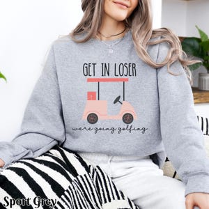 K&ouml;nnte beinhalten: Ein grauer Pullover mit dem Text "Get in loser, we're going golfing" und einer pinkfarbenen Golfwagen-Grafik.