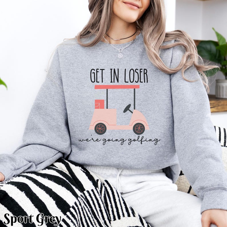 K&ouml;nnte beinhalten: Ein grauer Pullover mit dem Text "Get in loser, we're going golfing" und einer pinkfarbenen Golfwagen-Grafik.