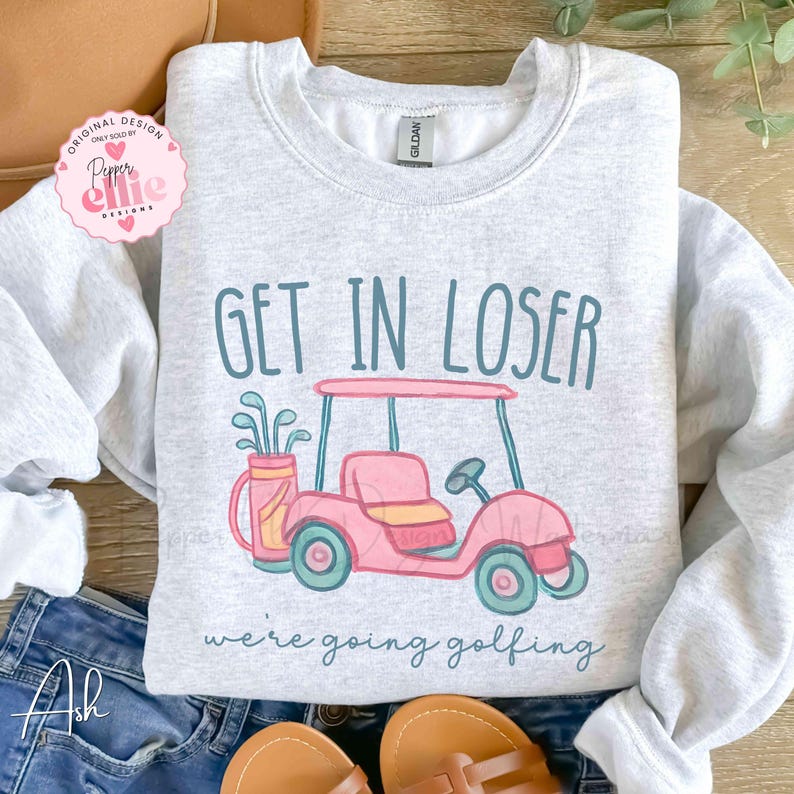 Puede incluir: Sudadera gris claro con el texto "GET IN LOSER" sobre un gr&aacute;fico de carrito de golf rosa. La frase "we're going golfing" est&aacute; impresa debajo. El dise&ntilde;o incluye una bolsa de golf con palos.