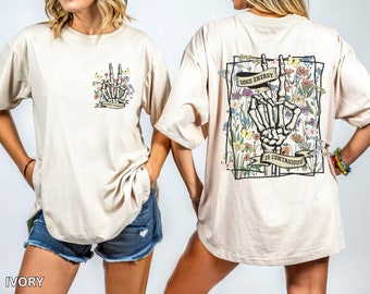 May include: A beige oversized t-shirt with a graphic print of a skeleton hand making a peace sign surrounded by flowers. The text "Good Energy is Contagious" is printed on a banner below the hand. The shirt is available in ivory.