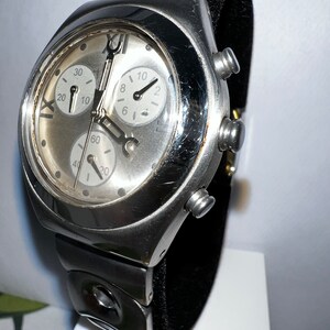 May include: A silver-toned wristwatch with a black band. The watch face has three sub-dials and Roman numeral markings.
