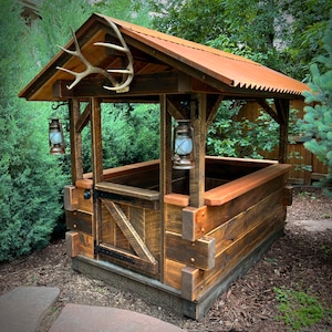 May include: A rustic wooden structure with a metal roof, featuring a large antler mounted above the entrance. Two antique-style lanterns hang on either side of the entrance. The structure is built with reclaimed wood and has a weathered, rustic look.