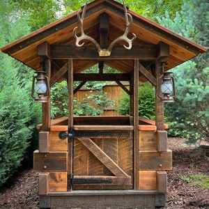 May include: A rustic wooden structure with a gabled roof and a wooden door. The structure is decorated with a pair of antlers and two lanterns. The structure is surrounded by green foliage.