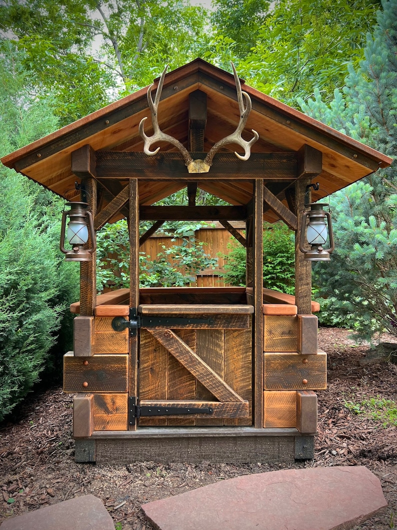 May include: A rustic wooden structure with a gabled roof and a wooden door. The structure is decorated with a pair of antlers and two lanterns. The structure is surrounded by green foliage.