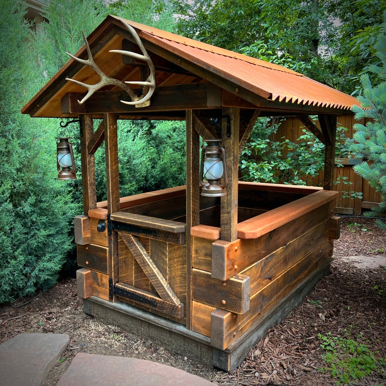May include: A rustic wooden structure with a metal roof, featuring a large antler mounted above the entrance. Two antique-style lanterns hang on either side of the entrance. The structure is built with reclaimed wood and has a weathered, rustic look.