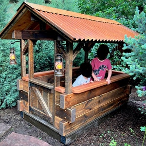 May include: A wooden playhouse with a corrugated metal roof, two hanging lanterns, and a wooden fence. Two children are playing inside the playhouse.