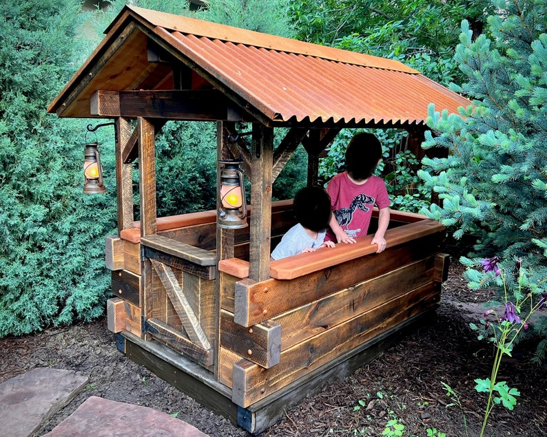 May include: A wooden playhouse with a corrugated metal roof, two hanging lanterns, and a wooden fence. Two children are playing inside the playhouse.