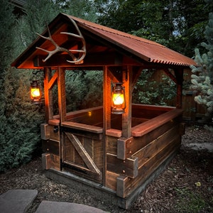 May include: A rustic wooden structure with a metal roof, featuring a large antler mounted above the entrance. Two lit lanterns hang on the sides, illuminating the interior. The structure has a wooden door and a wooden bench.