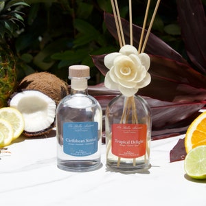 May include: Two glass bottles of reed diffusers with wooden sticks. The bottle on the left is blue and says "Caribbean Santal". The bottle on the right is orange and says "Tropical Delight". Both bottles are on a white surface.