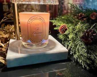 May include: A scented candle in a glass jar with the text "Tis Christmas" sits on a marble base, under a glass lamp. The candle has a peach hue and is surrounded by greenery and pinecones, creating a festive atmosphere.