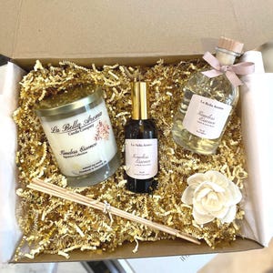 May include: A gift box filled with gold-colored shredded paper, containing a candle, a fragrance spray, a diffuser bottle, a flower, and reed sticks. The products are labeled with "La Bella Aroma" and "Timeless Essence".