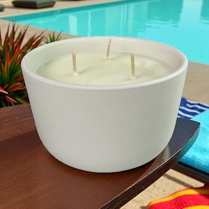 May include: A large, white, round candle with three wicks. The candle sits on a dark brown wooden tray, with a swimming pool in the background. The candle is unscented and has a smooth, matte finish.
