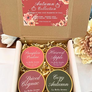 May include: An open cardboard box containing four scented candles from the La Bella Aroma Autumn Collection. The candles are in gold tins with labels that read "Pumpkin Praline", "Harvest Apple", "Spiced Cognac", and "Cozy Autumn". The box also has a decorative floral design.