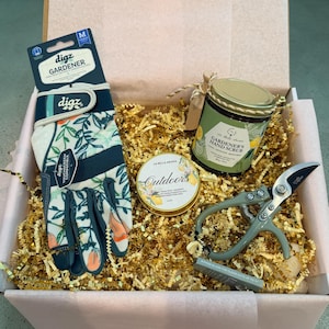 May include: A gift box containing gardening gloves, hand scrub, a tin of Outdoors balm, and pruning shears. The gloves are floral patterned with the brand name "digz". The hand scrub is in a green jar with a gold lid. The shears are gray.