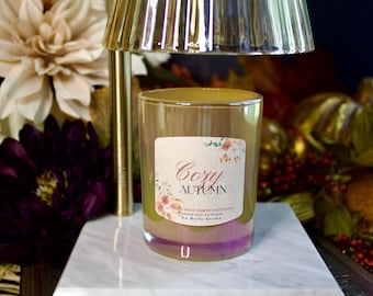 May include: A glass candle with a label that reads "Cozy Autumn" with floral accents. The candle has a gold and purple iridescent finish. A gold lamp and flowers are in the background. The candle is on a white marble surface.