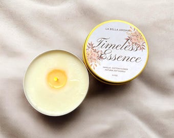 May include: A vanilla and cotton flower scented natural soy candle in a silver tin. The candle is labeled "Timeless Essence" with floral accents. The tin is 2.5 oz. and has a gold rim. The candle is lit, showing a yellow flame.