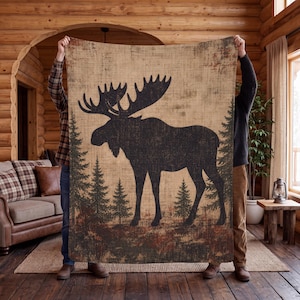May include: A rectangular throw blanket featuring a large black silhouette of a moose standing in a forest of pine trees. The background has a textured, tan, and brown weathered appearance, creating a rustic look across the entire fabric surface.