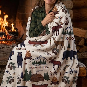 May include: A cream-colored fleece blanket featuring a repeating pattern of outdoor wilderness motifs including black bears, moose, deer, pine trees, log cabins, campfires, canoes, fishing rods, lanterns, and pinecones with geometric accents.