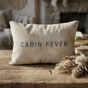 May include: A rectangular beige pillow with the words "CABIN FEVER" printed in dark blue. The pillow is on a rustic wooden table, with dried plants and a book nearby. The background includes a fireplace and other home decor.