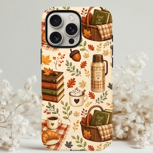 May include: A cream-colored phone case featuring a repeating autumn-themed pattern of brown wicker baskets, stacks of books labeled Leaf & Lore, plaid thermoses, mugs with heart designs, acorns, croissants, and colorful fall leaves.