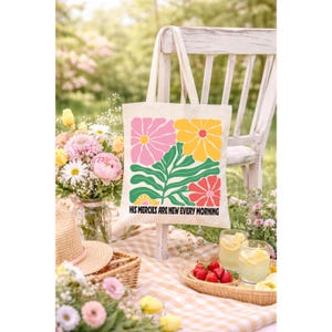 May include: Cream-colored tote bag with a floral design in pink, yellow, and red, with green leaves. The text "HIS MERCIES ARE NEW EVERY MORNING" is printed at the bottom. The bag is hanging on a white wooden chair.