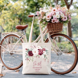 Könnte beinhalten: Eine cremefarbene Tragetasche mit einem Blumendesign aus bordeauxroten und weißen Blumen und dem Text "Be still and know that I am God" vor einem Vintage-Fahrrad mit einem Blumenkorb.