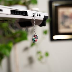 May include: A gray and red mushroom charm hangs from a silver chain attached to a white laptop.