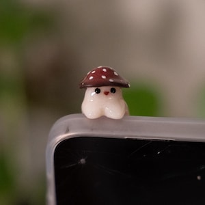 May include: A whimsical mushroom figurine with a brown cap and white polka dots. The mushroom has a cream-colored body and a smiling face. The figurine is small and appears to be made of ceramic or resin.