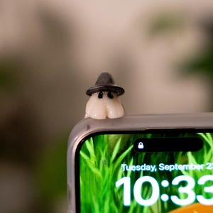 May include: A small, white ghost figurine wearing a black witch hat, perched on the edge of a phone. The phone screen displays a green background with the time 10:33 and the date Tuesday, September 23.