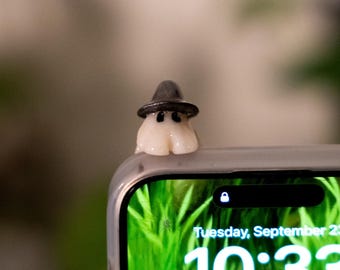 May include: A small, white ghost figurine wearing a black witch hat, perched on the edge of a phone. The phone screen displays a green background with the time 10:33 and the date Tuesday, September 23.