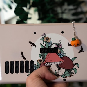 May include: A pink and black Nintendo Switch console with a custom design featuring a mushroom, flowers, and the word "VALVE". A small orange pumpkin charm hangs from the top of the console.