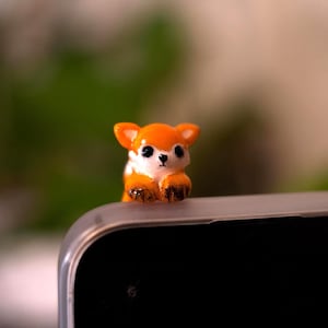 Peut inclure: Une petite figurine de renard orange et blanc brillant avec des yeux et un nez noirs, perchée sur le bord supérieur d'un écran sombre. Le renard a des oreilles pointues et des pattes aux extrémités sombres posées sur la surface.