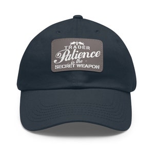 Puede incluir: Una gorra de béisbol azul marino con un parche gris que dice "TRADER Patience is the SECRET WEAPON".