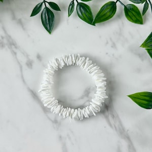 Puede incluir: Pulsera de conchas blancas hecha de pequeñas piezas planas de concha blanca. La pulsera está colocada sobre una superficie de mármol blanco.