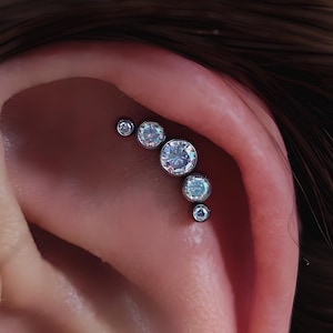 May include: Close-up of an ear with multiple piercings. Each piercing features a small, round, clear gemstone. The gemstones are set in silver-colored metal and are arranged in a curved line along the ear's cartilage.