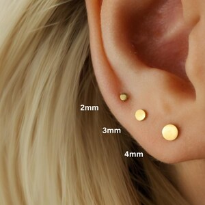 May include: Close-up of an ear with three gold-coloured disc stud earrings. The earrings are 2mm, 3mm, and 4mm in diameter. The ear is next to blonde hair. The text on the image reads "2mm", "3mm", and "4mm".