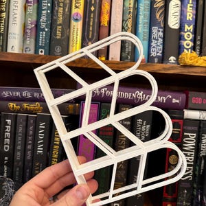 May include: A white, geometric letter "B" bookshelf, held in front of a bookshelf filled with books. The letter is constructed with a series of straight lines and curves, creating a modern, open design. The books have various titles and colors.